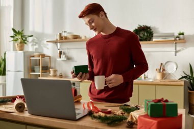 With a warm smile, a young man holds a gift while sipping a drink in a festive kitchen. clipart