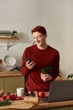 Hediye taşıyan neşeli genç bir adam, mutfaktaki telefonunu kontrol ederken gülümsüyor..