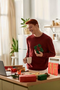 A young man smiles while holding a gift, enjoying his time in a cheerful and festive kitchen. clipart