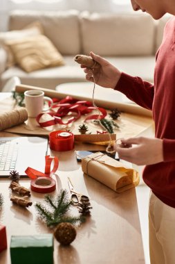 Genç bir adam Noel hediyelerini paketliyor, etrafı şenlikli dekorasyon ve malzemelerle çevrili..