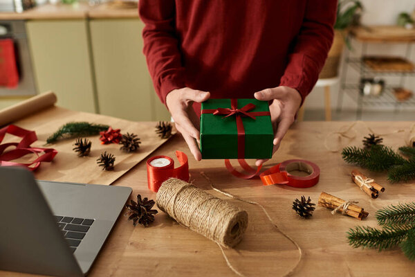 A young man wraps a green gift box with a red ribbon while celebrating Christmas at home.