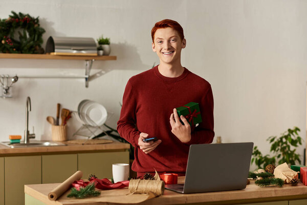 A young man with red hair is smiling as he holds a gift while surrounded by Christmas decorations.