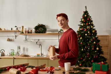 Neşeli genç bir adam sıcak bir mutfakta duruyor, süslenmiş bir Noel ağacının yanında hediyeleri paketliyor..