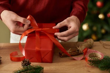 Genç bir adam gülümserken güzel paketlenmiş bir Noel hediyesinin etrafına canlı bir kurdele bağlıyor..