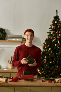 A cheerful young man stands by a decorated kitchen counter, holding a wrapped gift. clipart