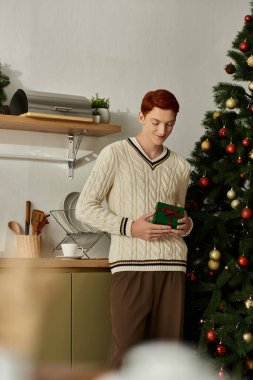 Genç bir adam elinde bir hediyeyle gülümsüyor, etrafı şenlikli süslemeler ve bir Noel ağacıyla çevrili..