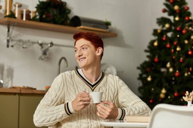 Genç bir adam elinde bir kupa tutarken gülümsüyor, etrafı şenlikli Noel süsleriyle çevrili..