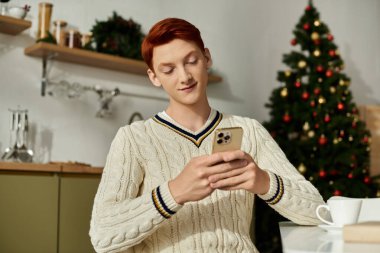 A young man with ginger hair smiles as he relaxes and checks his phone amidst festive decorations. clipart