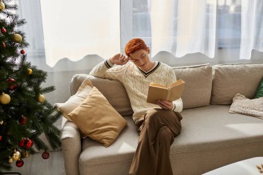 A young man sits comfortably on the sofa, immersed in a book while celebrating Christmas. clipart