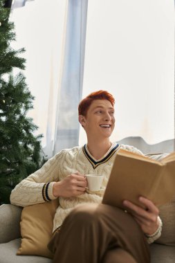 Neşeli genç bir adam Noel ağacının yanında rahatlar, sıcak bir içki içer ve kitap okur..