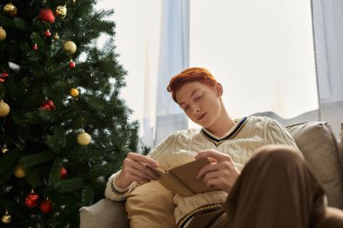 Genç bir adam güzel süslenmiş bir Noel ağacının yanında bir kitapla rahatlar..