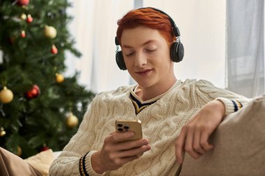 Genç adam süslü bir Noel ağacının yanında telefonunu kullanarak gülümsüyor..