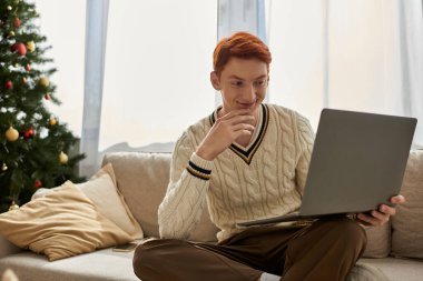 Genç bir adam kanepeye uzanır, dizüstü bilgisayarına gülümser, etrafı tatil dekoruyla çevrilidir..