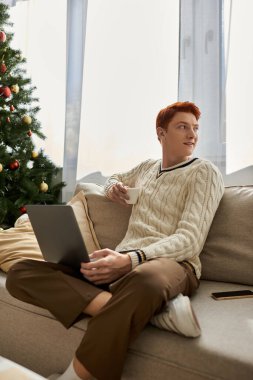 This young man relaxes on a sofa, sipping coffee while focused on his laptop during Christmas. clipart