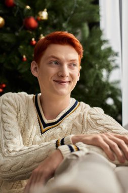Kızıl saçlı genç bir adam güzel süslenmiş bir Noel ağacının yanında gülümsüyor..