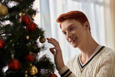 Neşeli bir genç adam güzel dekore edilmiş bir Noel ağacına renkli süsler ekliyor..