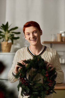 Neşeli genç bir adam, böğürtlenlerle süslenmiş bir Noel çelengini sunarken gülümsüyor..