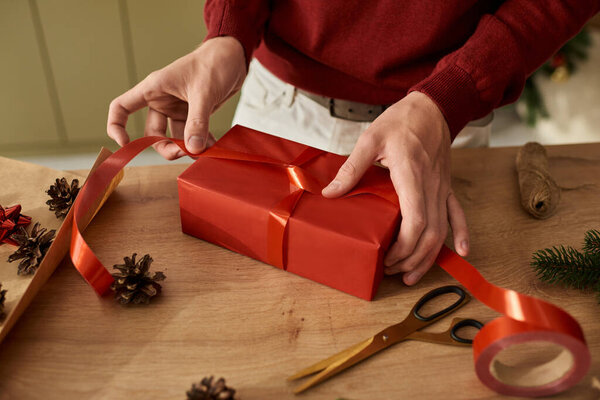 Молодой человек тщательно обертывает праздничный подарок яркой красной ленточкой, источая праздничное веселье.