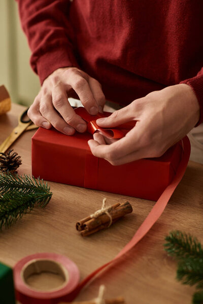 Молодой человек аккуратно завязывает ленточку вокруг ярко-красного подарка во время подготовки к Рождеству.