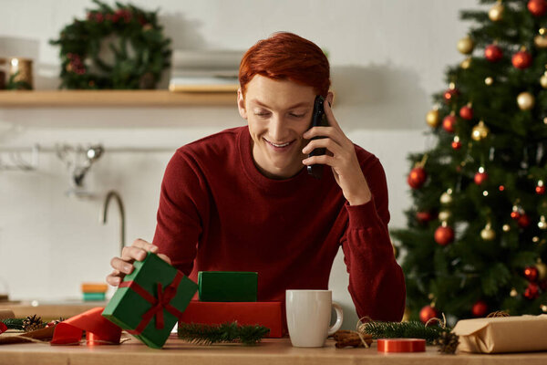 Молодой человек улыбается, разговаривая по телефону, взволнованно открывая рождественские подарки дома.