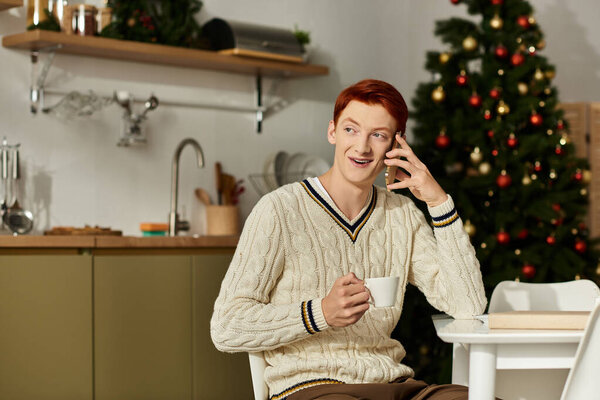 In a cozy kitchen, a young man smiles as he talks on the phone, holding a cup of coffee.