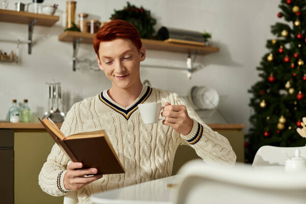 A joyful young man relaxes with a book in hand, savoring a warm drink during the holiday season.