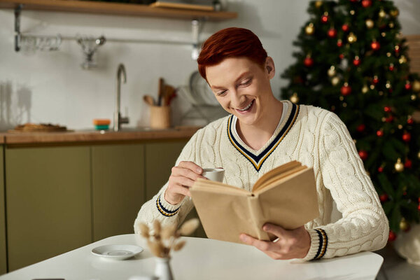 The young man smiles while sipping coffee and reading a book beside a decorated Christmas tree.