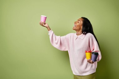 Genç bir kadın geniş bir alanda gülümsüyor. Renkli kahve fincanlarını yeşil bir arka planla dengeliyor..