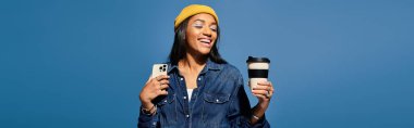 A cheerful young woman, dressed in autumn layers, holds a coffee cup and phone, radiating happiness. clipart