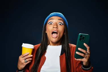 Sıcak sonbahar kıyafetleri içindeki genç bir kadın elinde kahve ve akıllı telefonuyla neşeyle tepki veriyor..