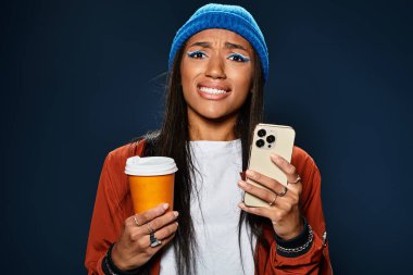 Sonbahar modasına uygun giyinmiş genç bir kadın içki ve telefon tutarken karışık duygular ifade ediyor..