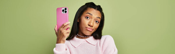 Стильная молодая женщина с игривым выражением лица держит розовый телефон, показывая яркие осенние вибрации.