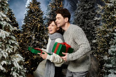 A smiling couple holding colorful gifts enjoys a magical winter moment amongst snowy fir trees. clipart