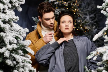 Kışlık ceketli romantik bir çift, karla kaplı çam ağaçlarıyla çevrili neşeli bir anın tadını çıkarıyor..