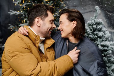 Sevgi dolu bir çift, karla kaplı çam ağaçlarıyla çevrili Noel neşesini kutlarken birbirlerine sıcak bir şekilde gülümserler..