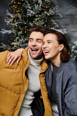 A happy couple embraces, wearing cozy jackets, surrounded by snow-covered trees, celebrating the Christmas spirit. clipart