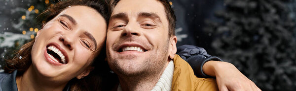 Счастливая пара обнимает дух Рождества в теплых куртках в окружении заснеженных сосен.