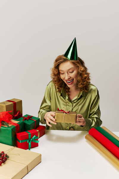 Молодая женщина, завернутая в зеленую рубашку, радостно держит подарки, надевая шляпу для вечеринок..