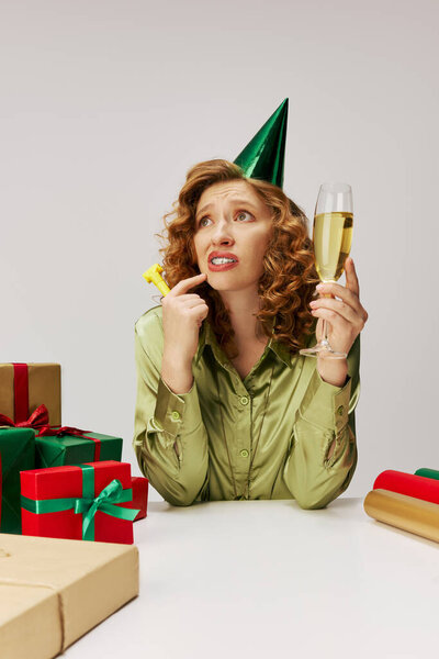 A cheerful young woman posing, holding a glass of sparkling drink and wearing a party hat.