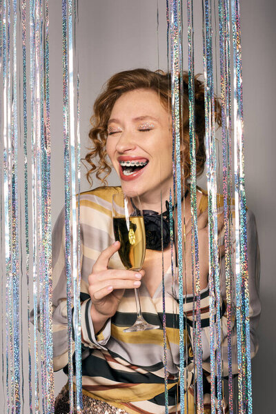 A cheerful young woman enjoys a drink amidst colorful and shiny decorations.