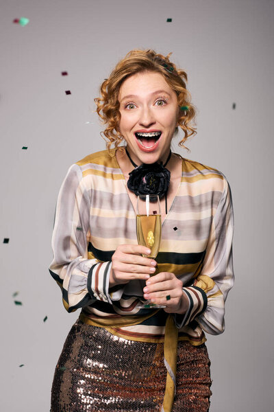 A happy young woman holds a champagne glass, surrounded by colorful confetti at a celebration.