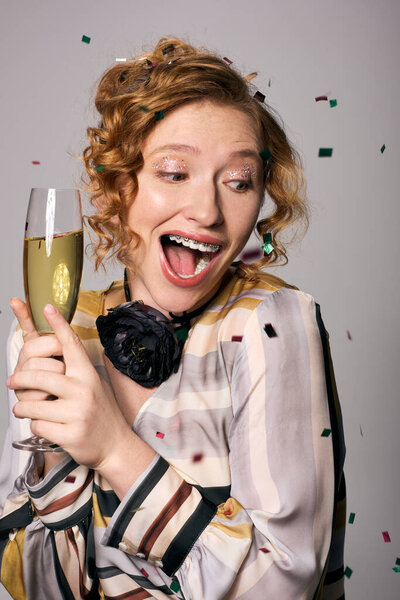 Возбужденная молодая женщина держит бокал шампанского и улыбается среди красочных конфетти и подарков.