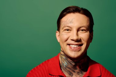 A tattooed young man grinning cheerfully in front of a bright green backdrop. clipart