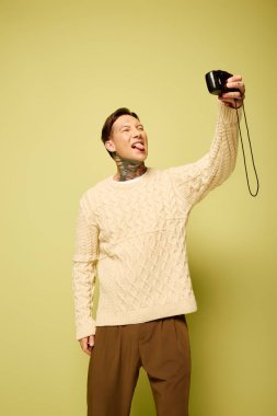 Dövmeleri olan genç bir adam fotoğraf makinesiyle selfie çekerken neşeli pozlar veriyor..