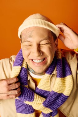 Dövmeli neşeli bir genç adam sırıtırken örgü bir şapka ve çizgili bir eşarp takıyor..