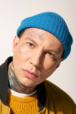 Çarpıcı dövmeleri olan genç bir adam mavi bir bere takmış kameraya bakıyor..