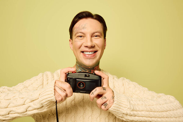 A cheerful young man displays a vintage camera and his unique tattoos in a lively setting.