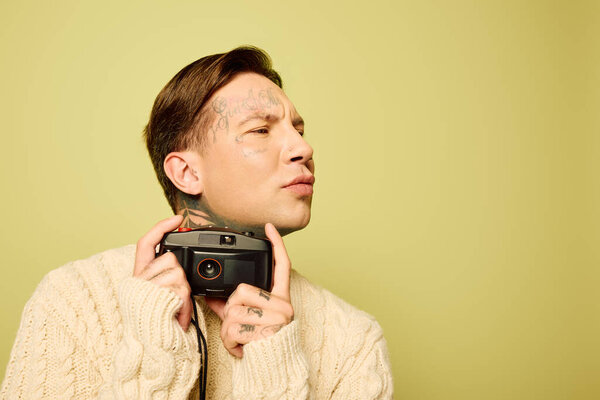 A stylish young man with intricate tattoos contemplates as he poses with a camera.