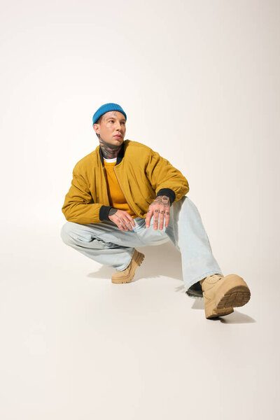 A young man with numerous tattoos squats confidently while showcasing a trendy outfit.