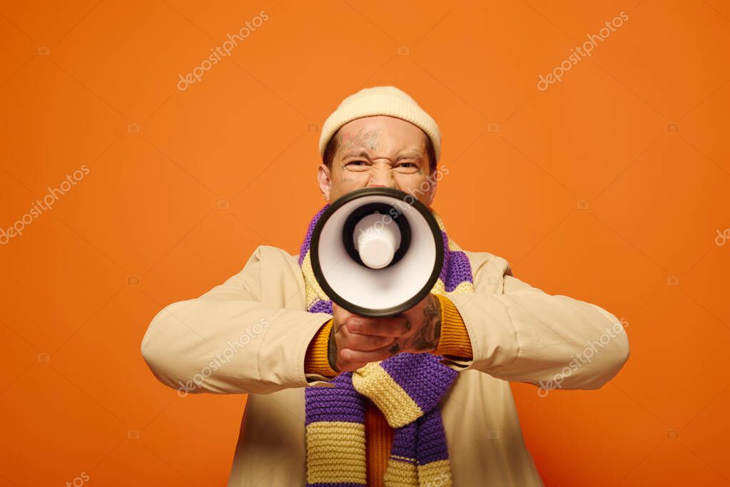 A young man with tattoos enthusiastically uses a megaphone against a bright orange backdrop.